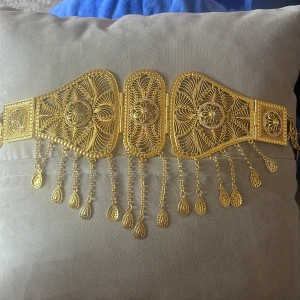 Kurdisk guld bälte 24k gold  - Unikt guldfärgat bälte med filigranmönster och hängande droppformade detaljer. Bältet har flera runda myntlänkar längs sidorna och ett dekorativt spänne. Perfekt för dig som vill sticka ut med en riktigt snygg accessoar. PRIS KAN DISKUTERAS 