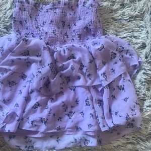 Ljuslila blommig kjol - Supersöt ljuslila kjol med smockad överdel och volangig kjol. Kjolen har ett blommigt mönster med små blommor i blått och gult. Perfekt för sommarens alla tillfällen och ger en romantisk vibe.