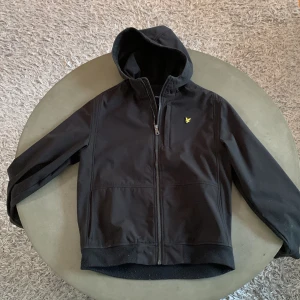 Svart vindjacka från Lyle & Scott - Svart vindjacka från Lyle & Scott med huva och dragkedja framtill. Jackan har en stilren look med gul logga på bröstet och är tillverkad i ett slätt syntetmaterial. Perfekt för blåsiga dagar och enkel att matcha med det mesta.