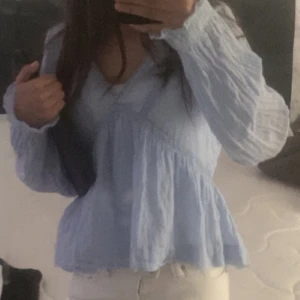 Ljusblå blus med v-ringning storlek 164-170! - Säljer en ljusblå blus med v-ringad hals och volang nedtill. Blusen har långa ärmar och är tillverkad i ett tunt, luftigt bomullstyg. Snygga sömmar och lätt rynkade detaljer ger en chill och feminin vibe. Perfekt att styla med jeans eller kjol. Råkat dra bort Gina märket!!! Storlek 164 till 170!