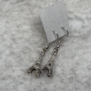 Örhängen med Eiffeltorn i silver - Snygga örhängen med hängande Eiffeltorn i silverfärgad metall. Perfekt för dig som älskar Paris eller vill ha en unik detalj till din stil. De har krokfästen och är lätta att bära. Ge din look en touch av äventyr och romantik!