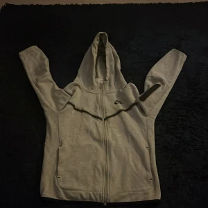Nike tech fleece 147-158 - Säljer en grå hoodie från Nike med hel dragkedja och huva. Tröjan har svarta detaljer, långa ärmar och en sportig look. Perfekt för chill dagar eller när du vill ha en enkel och clean stil. Passar dig som gillar streetwear och bekväma plagg.