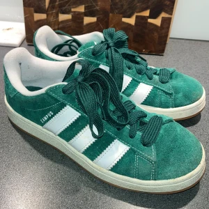 Adidas Campus gröna sneakers - Gröna Adidas Campus sneakers med klassiska vita ränder på sidorna och gröna snören. Ovandel i mjuk mocka och vit insida, platt sula och rund tå. Ikonisk Adidas-logga, storlek 41 1/3, Urpsungspris: 1100kr Nytt pris: 600kr