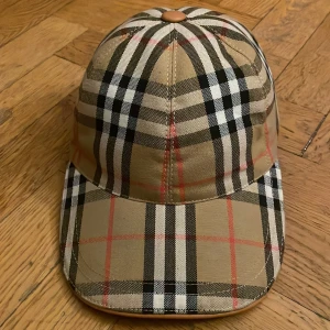 Burberry rutig keps  - Snygg keps från Burberry med klassiskt rutigt mönster i beige, svart, vitt och röda linjer. Justerbar rem baktill och böjd skärm. Tillverkad i bomull och perfekt för dig som vill ha en ikonisk accessoar med streetkänsla.