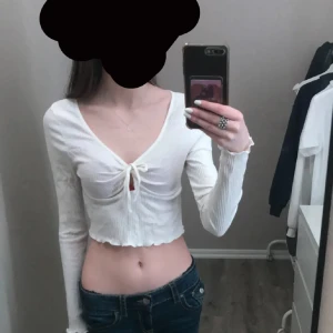 Vit croppad ribbad topp med knyt - Säljer en vit croppad topp med långa ärmar och v-ringning. Toppen har ribbad struktur och ett snyggt knyt framtill som ger en cool detalj. Materialet är mjukt och stretchigt, perfekt för dig som gillar trendiga och bekväma plagg.