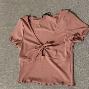 Rosa croppad topp från SHEIN - Supersöt ljusrosa croppad topp från SHEIN med ribbad struktur och korta ärmar. Toppen har v-ringning och en snygg knytning framtill. Ärmsluten och nederkanten har volangdetaljer som ger en extra touch. Perfekt för dig som gillar trendiga och feminina plagg.ny oanvänd eller använd 1 gång.
