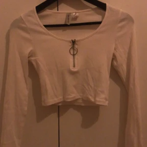 Vit croppad långärmad topp H&M - Säljer en vit croppad långärmad topp från H&M Divided i ribbad bomull. Toppen har rund hals och en cool dragkedja med stor ring framtill. Perfekt för dig som gillar stilrena och trendiga detaljer.