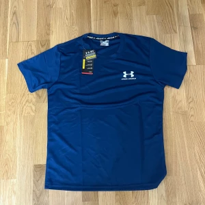 Blå kompressionst-shirt från Under Armour(helt ny och gratis frakt) - Snygg marinblå kompressionst-shirt från Under Armour med vit logga på bröstet och nacken. Tillverkad i ett lätt och snabbtorkande syntetmaterial, perfekt för träning. Klassisk rund hals och korta ärmar. Sitter tight och ger en sportig look.