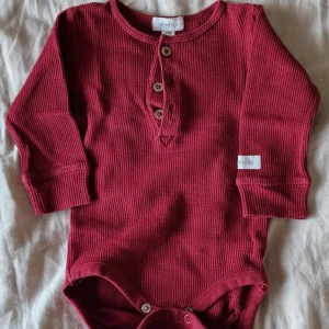 Vinröd body från Newbie - Vinröd långärmad body från Newbie i bomull med knappar framtill. Perfekt för höst och vinter. Snygg och enkel design med rund hals och praktiska tryckknappar i grenen.