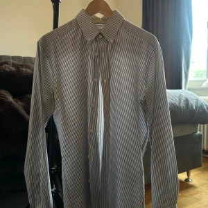 Randig skjorta These Glory Days - Snygg skjorta från These Glory Days i slim fit-modell. Skjortan är vit med smala blå ränder, har klassisk krage med knappar och långa ärmar. Perfekt för dig som gillar stilren och skandinavisk design.