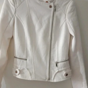 Vit bikerjacka med dragkedjor - Snygg vit bikerjacka med asymmetrisk dragkedja och två fickor med dragkedjor. Jackan har långa ärmar, markerad midja och coola knappdetaljer vid höften. Materialet är strukturerat och ger en fräsch look.