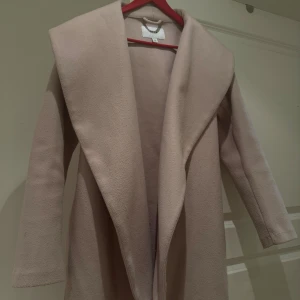 Beige omlottkappa från VILA - Snygg beige kappa från VILA med bred krage och omlottmodell. Jackan har ett bälte i midjan för att knyta och långa ärmar. Materialet är mjukt och känns som ullblandning, perfekt för kyliga dagar. Enkel och stilren design som ger en clean look.