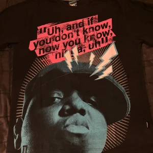 Svart t-shirt med tryck Notorious B.I.G. - Svart t-shirt från the Notorious B.I.G / Biggie Smalls. med stort grafiskt tryck framtill i rött, blått och vitt. Texten är tryckt i en cool, streetinspirerad stil och motivet har blixtar och siluett. Klassisk passform och rund halsringning.