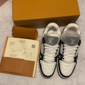 Louis Vuitton svartvita sneakers - Säljer ett par Louis Vuitton sneakers i svart och vitt med tydliga LV-loggor på tungan och sidan. Skorna har snörning, platt sula och är tillverkade i en mix av skinn och textil. Snygg kontrast mellan svart och vitt, samt diskreta monogramdetaljer på panelerna.