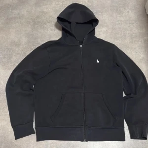 Svart hoodie från Polo Ralph Lauren - Svart hoodie från Polo Ralph Lauren med dragkedja och klassisk vit broderad logga på bröstet. Tröjan har huva, känguruficka och är tillverkad i mjuk bomullsmix. Perfekt för en chill och stilren look.