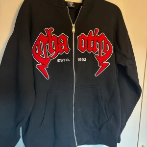Svart hoodie med röd text från FSBN - Svart hoodie från FSBN med dragkedja och stor röd text i grafisk stil på bröstet. Modellen har huva, kängurufickor och relaxed fit. Perfekt för dig som gillar streetwear och vill sticka ut med en cool detalj.