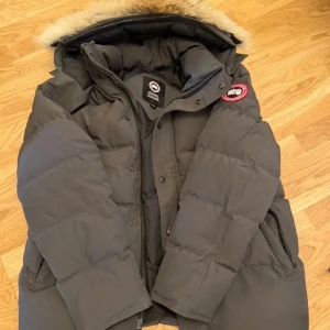 Grå dunjacka från Canada Goose M - Säljer en grå dunjacka från Canada Goose med pälskantad huva och klassisk logga på ärmen. Jackan är quiltad, har dragkedja och knappar framtill samt flera fickor. Perfekt för kalla vinterdagar och riktigt snygg passform.