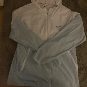 Nike vit och ljusblå vindjacka M - Nike vindjacka i vitt och ljusblått med dragkedja framtill och huva. Jackan har meshfoder, två fickor med dragkedja och klassisk Nike-logga på bröstet. Perfekt för blåsiga dagar och sportiga outfits.