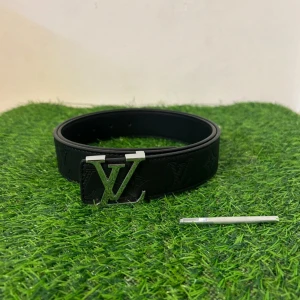 Louis Vuitton svart skärp med LV-spänne - Säljer ett svart Louis Vuitton bälte i skinn med ikoniskt LV-spänne i silver. Bältet har präglade LV-loggor och monogram längs hela remmen. Stilren och bred design som ger en lyxig vibe till outfiten. Perfekt för dig som vill ha något extra till jeansen.