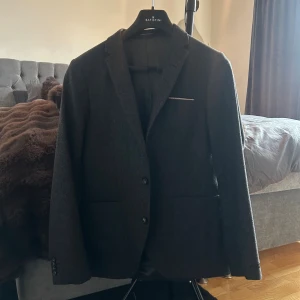 Mörkgrå kavaj från Tiger of Sweden - Snygg mörkgrå kavaj från Tiger of Sweden med klassisk passform. Kavajen har två knappar framtill, två fickor och en bröstficka med diskret detalj. Perfekt för dig som vill ha en stilren och tidlös look.