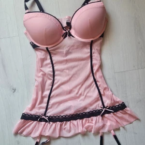 Rosa babydollklänning med svarta detaljer - Supersöt rosa babydollklänning med svarta spetsdetaljer och små rosetter. Klänningen har vadderade kupor, justerbara axelband och volangkant nertill. Materialet är mjukt och glansigt, perfekt för en extra feminin look.