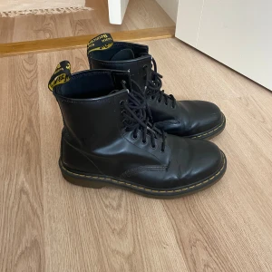 Dr. Martens svarta boots - Klassiska svarta Dr. Martens 1460 boots i skinn med snörning och ikonisk gul söm runt sulan. Grov platt sula och dragflik med logga baktill. Perfekta för dig som vill ha en tidlös och edgy stil.
