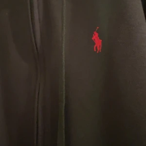 Svart hoodie från Polo Ralph Lauren - Svart hoodie från Polo Ralph Lauren med dragkedja framtill och röd broderad logga på bröstet. Tröjan har huva med snörning och känguruficka. Perfekt för en chill och stilren look.