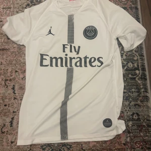 PSG vit kortärmad fotbollströja Jordan - Snygg vit PSG fotbollströja från Jordan med svart Paris-emblem och Fly Emirates-tryck på bröstet. Tröjan har en svart randig detalj i mitten och korta ärmar. Materialet är lätt och andas, perfekt för match eller träning. Röd och blå rand på insidan av kragen.