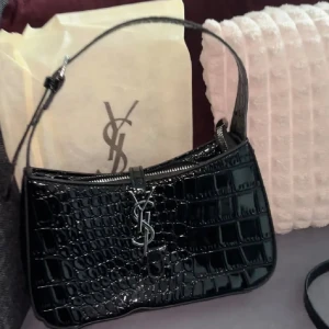 Svart axelväska - Snygg svart axelväska från med croco-mönstrad yta i glansigt skinn. Väskan har silverfärgad YSL-logga framtill och dragkedja upptill. Kommer med justerbar axelrem och dustbag med logga. 🫶🏻
