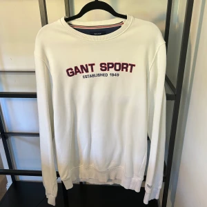 Vit sweatshirt från GANT med tryck - Vit sweatshirt från GANT med rött och svart broderat 'GANT SPORT' tryck på bröstet. Klassisk rund halsringning med ribbade muddar och nederkant. Perfekt för en sportig och clean stil.