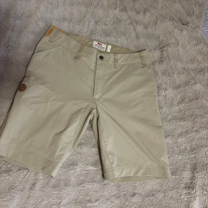 Beiga shorts från Fjällräven, strl 40 - OANVÄNDA!!!!!!!Snygga beiga shorts från Fjällräven i storlek 40. Klassisk modell med midwaist, bälteshällor och dragkedja bak. Perfekta för sommardagar och outdoor-häng. Materialet är slitstarkt och känns lätt på kroppen. Diskret logga på benet.