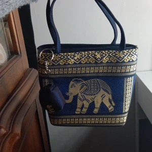 Blå och guld handväska med elefantmotiv - Unik handväska i mörkblått tyg med detaljer i guld och broderade elefanter. Väskan har två handtag, dragkedja upptill och ett matchande litet myntfodral som hänger i en nyckelring. Perfekt för dig som gillar färgstarka accessoarer med mönster.