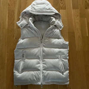 Vit dunväst från Philippe med huva - Snygg vit dunväst från Philippe i storlek M. Västen har en avtagbar huva, två fickor med dragkedja och quiltad design. Perfekt för lager-på-lager och håller dig varm under kyliga dagar. Stängs med dragkedja framtill och har en stilren look.