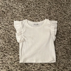Vit ribbad topp med volangärmar från Zara - Superfin vit ribbad topp från Zara med korta ärmar och volangdetaljer på axlarna. Toppen har rund halsringning och jätte gullig. Ganska kort i längden. Perfekt för dig som gillar en söt och stilren look.