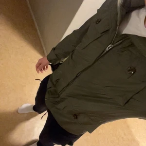 Grön vindjacka från J.Lindeberg - Snygg militärgrön vindjacka från J.Lindeberg med fyra stora fickor framtill och dragkedja hela vägen upp. Jackan har klassisk krage, axelklaffar och diskret logga på ärmen. Perfekt för dig som gillar stilren och funktionell design. 