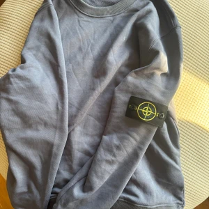 Grå sweatshirt från Stone Island - Säljer en stilren grå sweatshirt från Stone Island med klassisk logga på ärmen. Tröjan har rund hals, ribbade muddar och är tillverkad i mjuk bomull. Perfekt för dig som gillar streetwear och vill ha en clean look. Pris kan diskuteras 