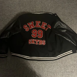 Svart bomberjacka med röd S och tryck - Svart bomberjacka från SKTBS med coola röda detaljer. Stor röd S på bröstet och SWEET 99 SKTBS tryckt på ryggen. Jackan har svarta ärmar i syntetmaterial, vita ränder på muddar och krage samt knappar framtill. Perfekt för dig som gillar streetstyle.