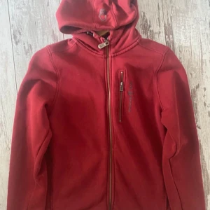 Röd hoodie från Sail Racing med dragkedja - Röd hoodie från Sail Racing med hel dragkedja framtill, huva med snörning och en bröstficka med dragkedja. Broderad logga på bröstet och mjukt material som känns skönt mot huden. Perfekt för kyliga dagar.
