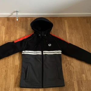 Sergio Tacchini softshelljacka med huva - Snygg softshell/vindjacka från Sergio Tacchini i svart och mörkgrått med röda detaljer på axlarna. Jackan har huva med snörning, dragkedja framtill, två fickor och vita ränder över bröstet. Perfekt för dig som gillar sportig stil.
