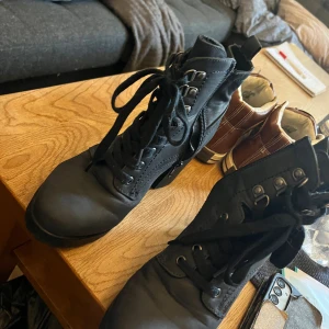 Svarta boots med klack och snörning - Säljer ett par svarta boots med grov klack, snörning framtill och dragkedja på sidan. Skorna har rund tå och är tillverkade i ett matt syntetmaterial. Perfekta för dig som vill ha en edgy look med stabilitet och höjd.