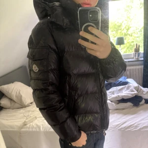 Svart Moncler pufferjacka med huva - Säljer denna riktigt feta moncler Maya i topp skick. Köpt på sp second place i Malmö som bara säljer äkta grejer o har kvitto, finns ett fel på sista bilden någon av knapparna är dåligt gjorde men funkar frf exakt lika bra som vanligt ser bara ut så.