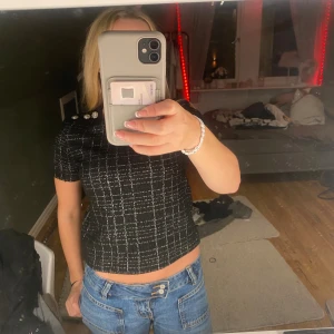 Svart rutig blus med silvriga knappar - Säljer en svart rutig blus från H&M med kort ärm och tre stora silvriga dekorationsknappar på ena axeln. Toppen har en boxig passform och är tillverkad i ett lite grövre tyg med vit tråd i rutmönster. Perfekt för dig som gillar en chic och trendig look.