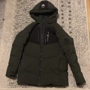 Grön Sail Racing dunjacka med huva - Mörkgrön och svart dunjacka från Sail Racing med huva, två bröstfickor med dragkedja och logga på bröstet. Jackan har en schysst pufferstil, justerbara ärmslut med kardborre och snygga svarta detaljer på axlar och huva. Jackan är lite sliten på få ställen, men annars är den i nyshick. Pris kan diskuteras för en snabb affär. Hör av er vid några frågor 👍