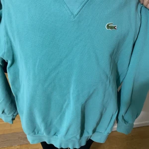 Turkos pikétröja från Lacoste - Snygg turkos pikétröja från Lacoste med klassisk krokodillogga på bröstet. Tröjan har v-ringad hals och långa ärmar med muddar. Materialet är bomull och har en typisk pikéstruktur som känns både sportig och chill.