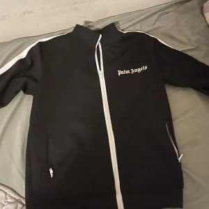 Svart track jacket från Palm Angels - Svart track jacket från Palm Angels med vita ränder längs ärmarna och vit dragkedja. Jackan har hög krage, två fickor med dragkedja och logga på bröstet. Perfekt för en sportig och trendig look.