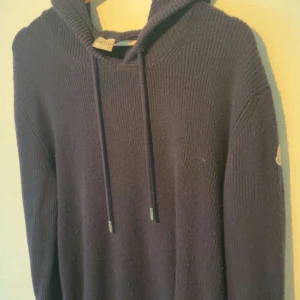 Mörkblå stickad hoodie från Moncler - Säljer en mörkblå stickad hoodie från Moncler med huva och snörning. Tröjan har långa ärmar och en klassisk Moncler-logga på ärmen. Perfekt för dig som gillar en chill och exklusiv stil. Jävkligt skön 