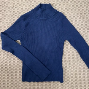 Marinblå ribbad polotröja - Säljer en marinblå ribbad polotröja med lång ärm och hög krage. Tröjan är stickad i ett mjukt material och har en slim passform som sitter snyggt på kroppen. Perfekt för lager-på-lager under kyliga dagar.