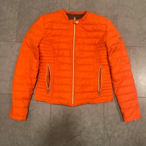 Orange pufferjacka från Rockandblue - Snygg orange pufferjacka från Rockandblue med markerade sömmar, dragkedjor på fickor och ärmslut samt coolt logomärke på ena ärmen. Jackan har en figurnära passform och är perfekt för dig som vill sticka ut med färg i höst och vinter.