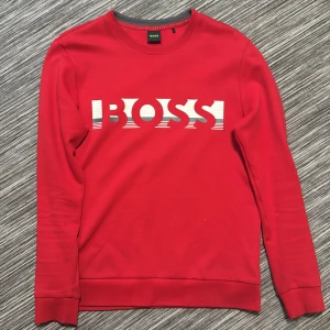 Röd BOSS sweatshirt med logga - Röd sweatshirt från BOSS med stor vit logga framtill i coolt grafiskt tryck. Tröjan har rund halsringning, långa ärmar och ribbade muddar. Tillverkad i mjuk bomull för en skön känsla och snygg passform.