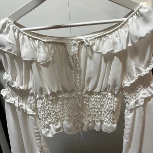 Vit offshoulder blus med volang - Supersöt vit offshoulder blus med volangdetaljer och smockad midja. Blusen har långa ärmar och knytning framtill. Perfekt för en romantisk look och passar till både jeans och kjol.